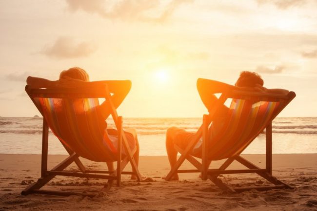 Romantic holiday travel. Silhouette of happy young couple sitting in deck chairs in luxury beach hotel at sunset near the sea. Love and relationship concept. Summer vacation in tropical paradise island.