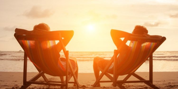 Romantic holiday travel. Silhouette of happy young couple sitting in deck chairs in luxury beach hotel at sunset near the sea. Love and relationship concept. Summer vacation in tropical paradise island.